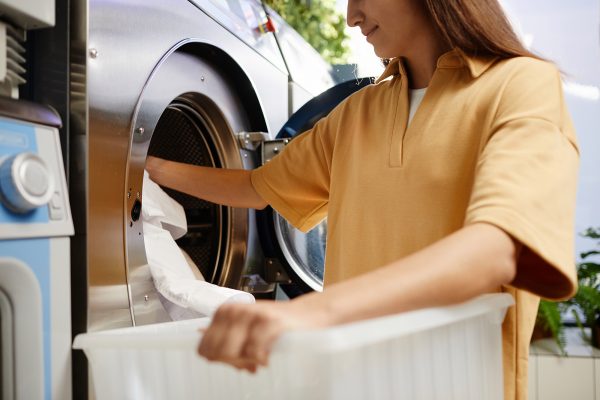 Girl in beige shirt putting white clothes into drum of washing machine from plastic container while standing in front of appliance